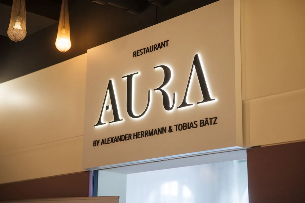 Beleuchtete Schriftzug-Tafel des Restaurants AURA über einem Durchgang, links zwei Hängelampen aus Metallgewebe