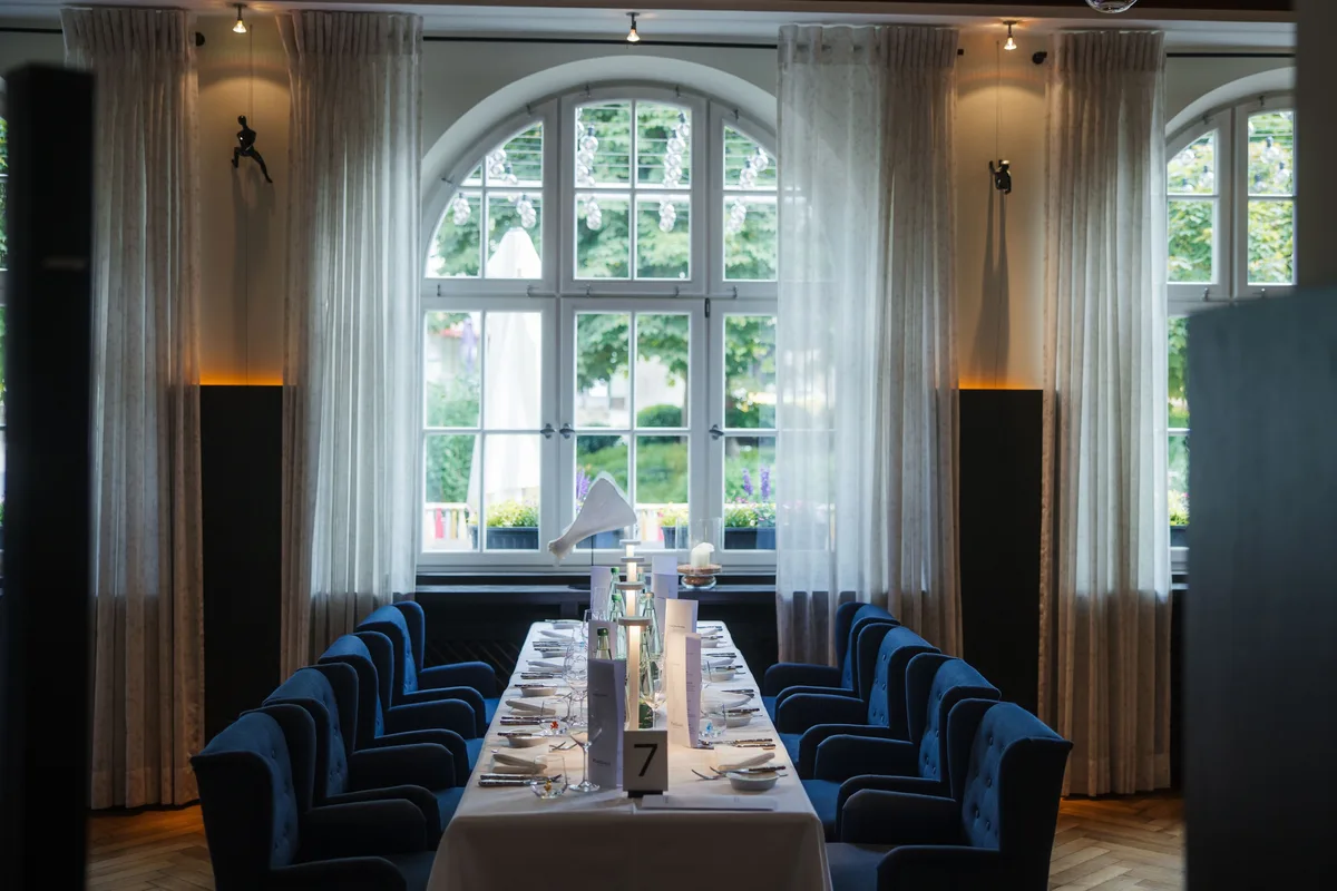 Lange gedeckte Tafel mit Nummer 7 vor einem Rundbogenfenster, beidseits blaue Polstersessel und helle Vorhänge