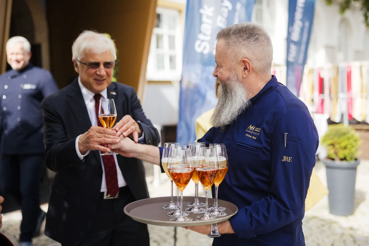Älterer Herr mit Brille nimmt von einem bärtigen Koch in blauer Jacke ein Glas vom Tablett, im Hintergrund blaue Fahnen