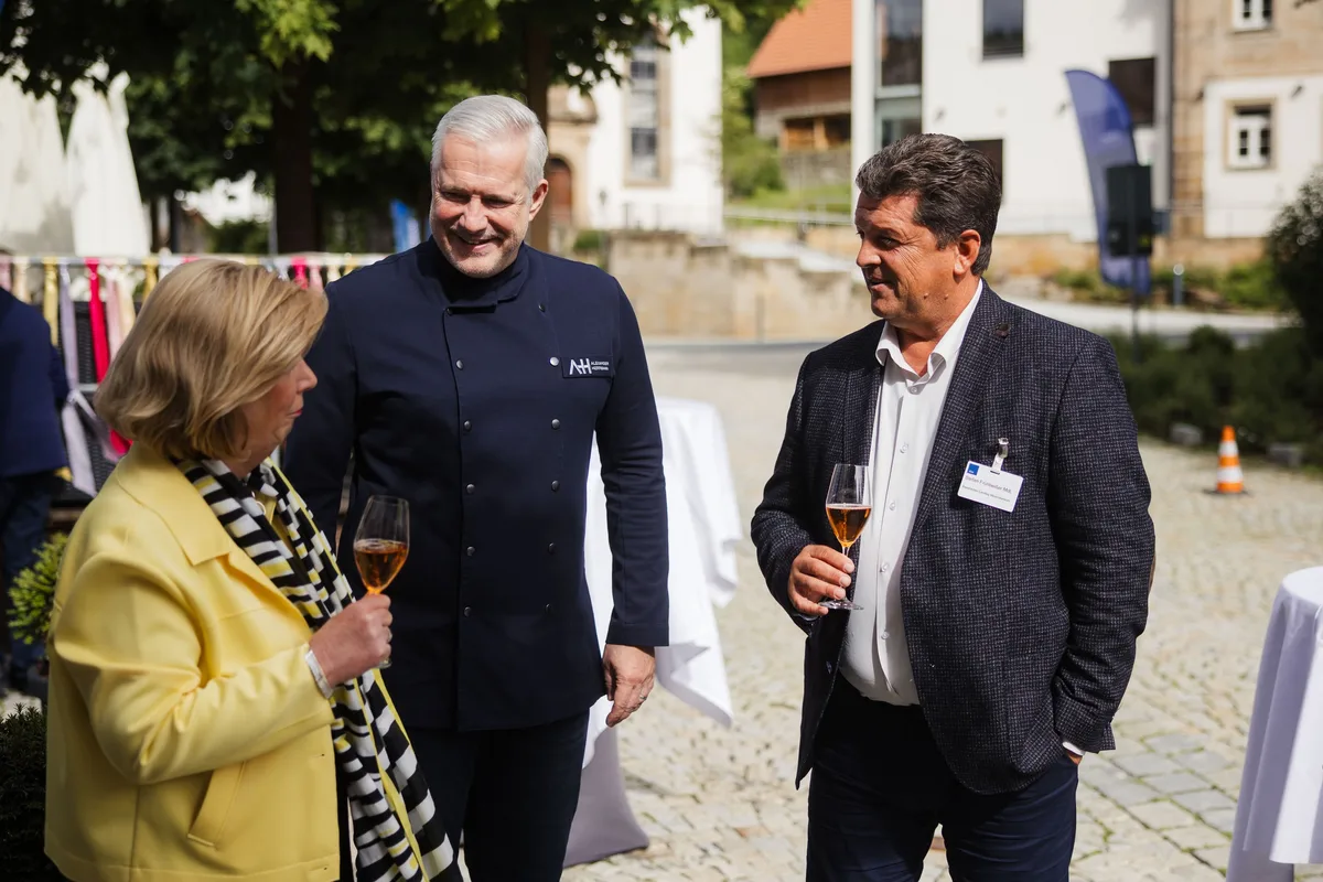 Frau in gelber Jacke, Koch in dunkler Jacke und Mann im Sakko stehen lachend mit Gläsern auf dem Pflaster zusammen