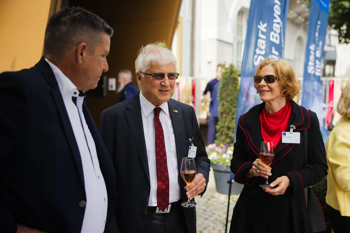 Zwei Männer und eine Frau mit Sonnenbrille und rotem Schal im Gespräch, alle mit Namensschild und Glas, dahinter blaue Fahnen