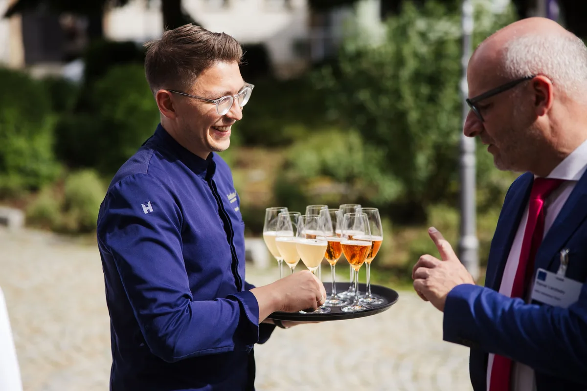Lachender Koch in blauer Jacke hält ein Tablett mit Gläsern, vor ihm ein Mann mit Brille und rotem Schlips im Anschnitt