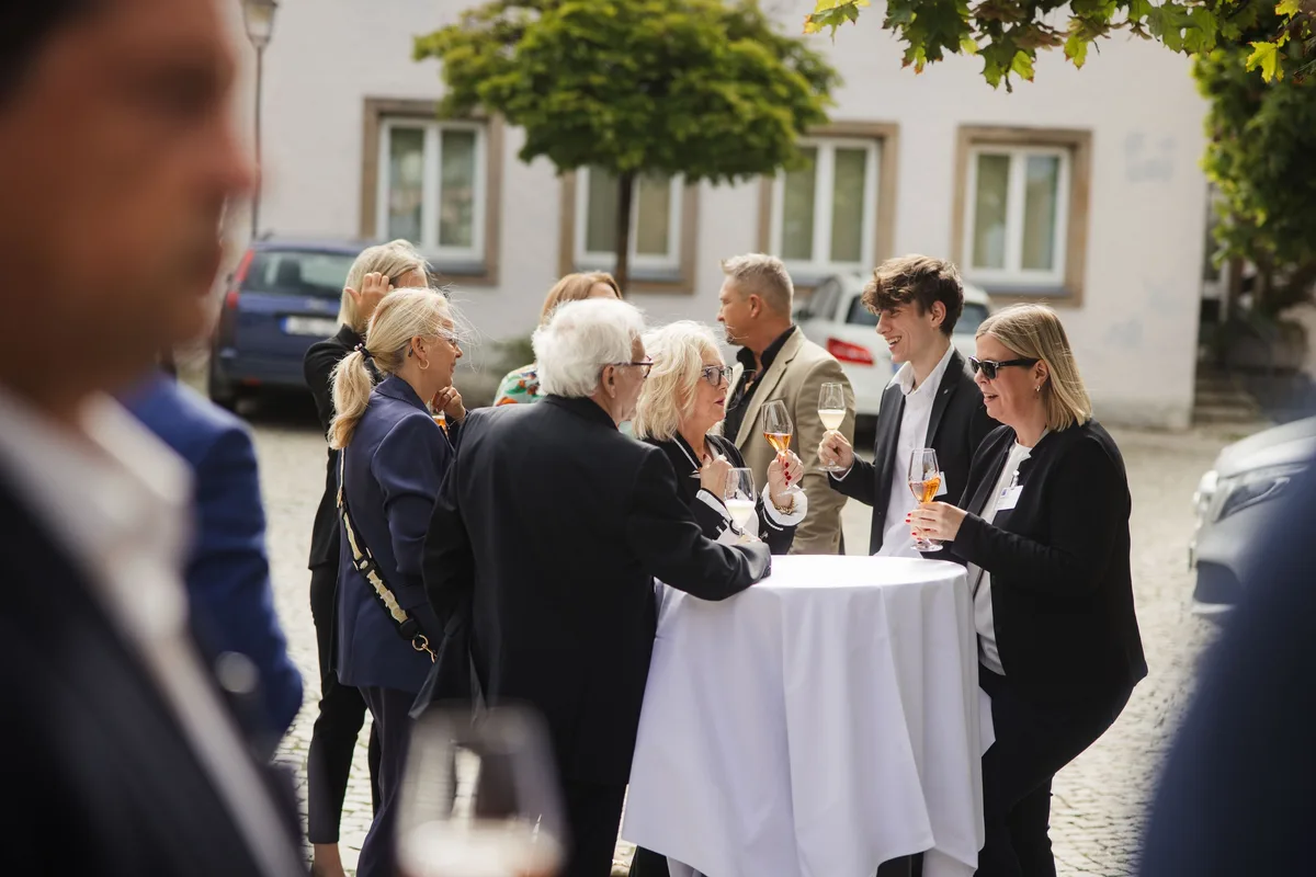 Gäste mit Gläsern stehen um einen Stehtisch mit weißem Tuch auf dem Marktplatz, im Vordergrund unscharfe Anschnitte