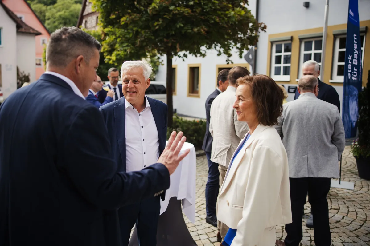 Mann im Sakko gestikuliert im Gespräch mit einem lachenden Mann in Hemd und Jackett, daneben eine Frau in heller Jacke