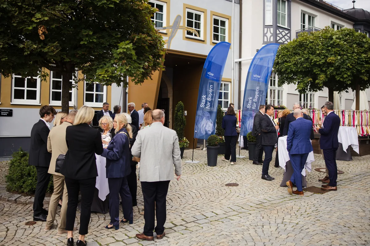Gäste stehen in Gruppen an Stehtischen vor dem Eingang des Posthotels, zwei blaue vbw-Fahnen, rechts bunte Stoffstreifen