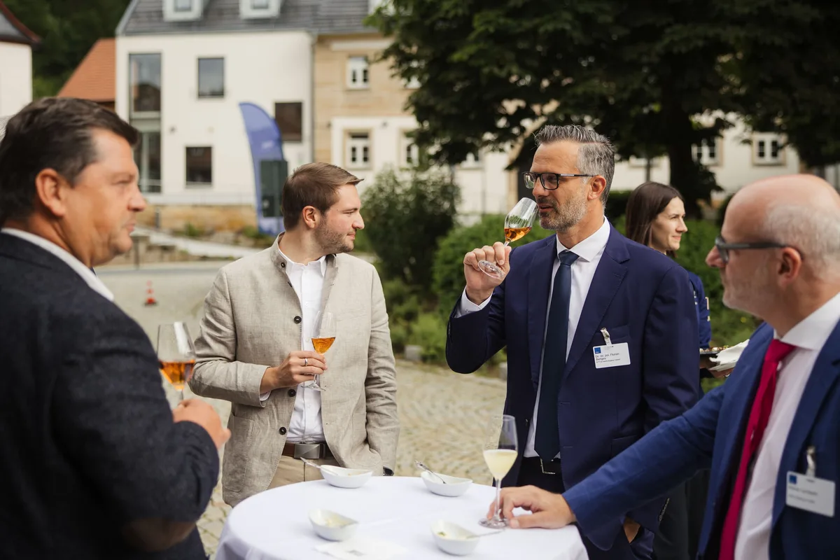 Zwei Männer trinken aus Gläsern am Stehtisch, rechts ein Mann mit Bart und Brille im Anzug, dahinter Marktplatz und Bäume