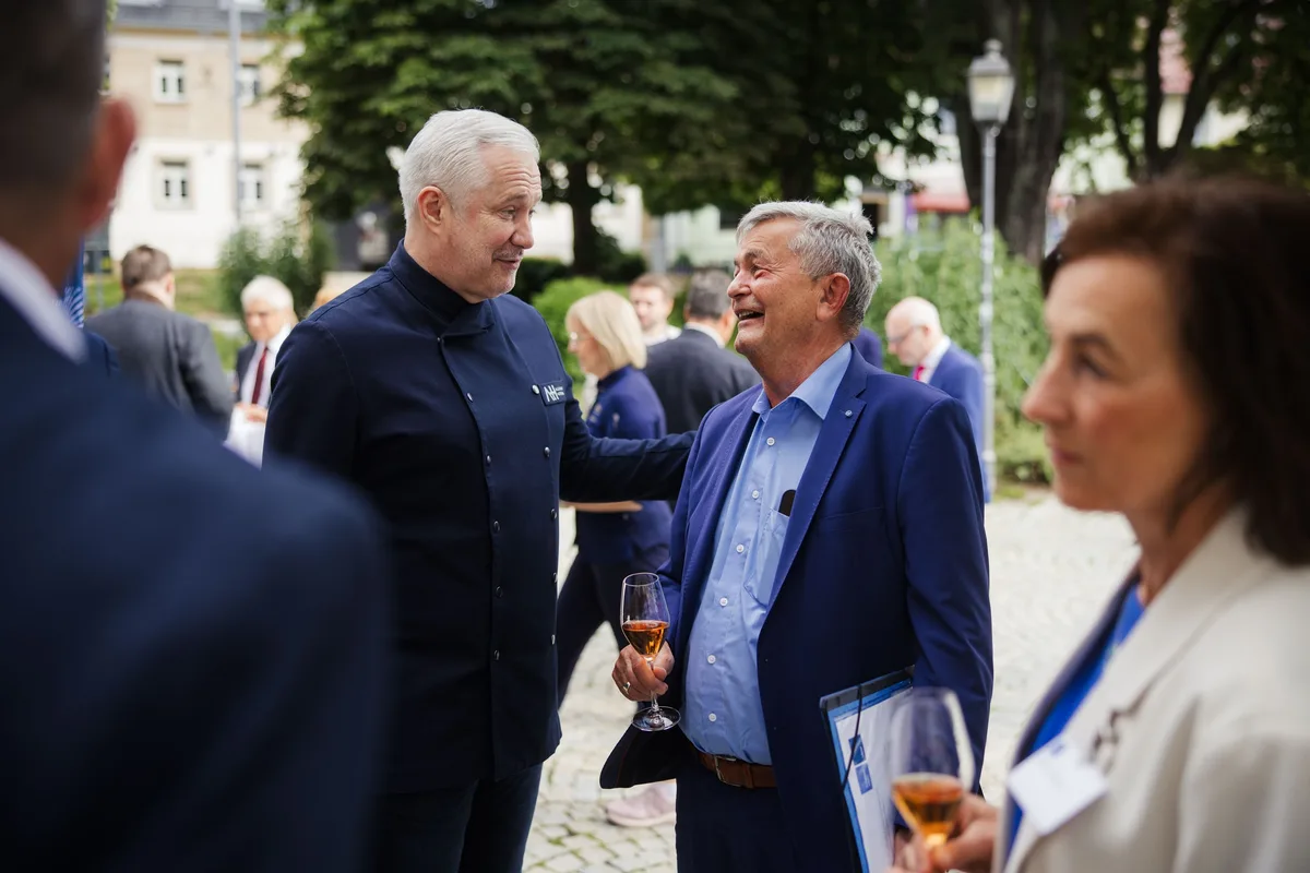 Koch in dunkler Jacke legt lachend die Hand auf die Schulter eines älteren Mannes im blauen Sakko, der ein Glas hält