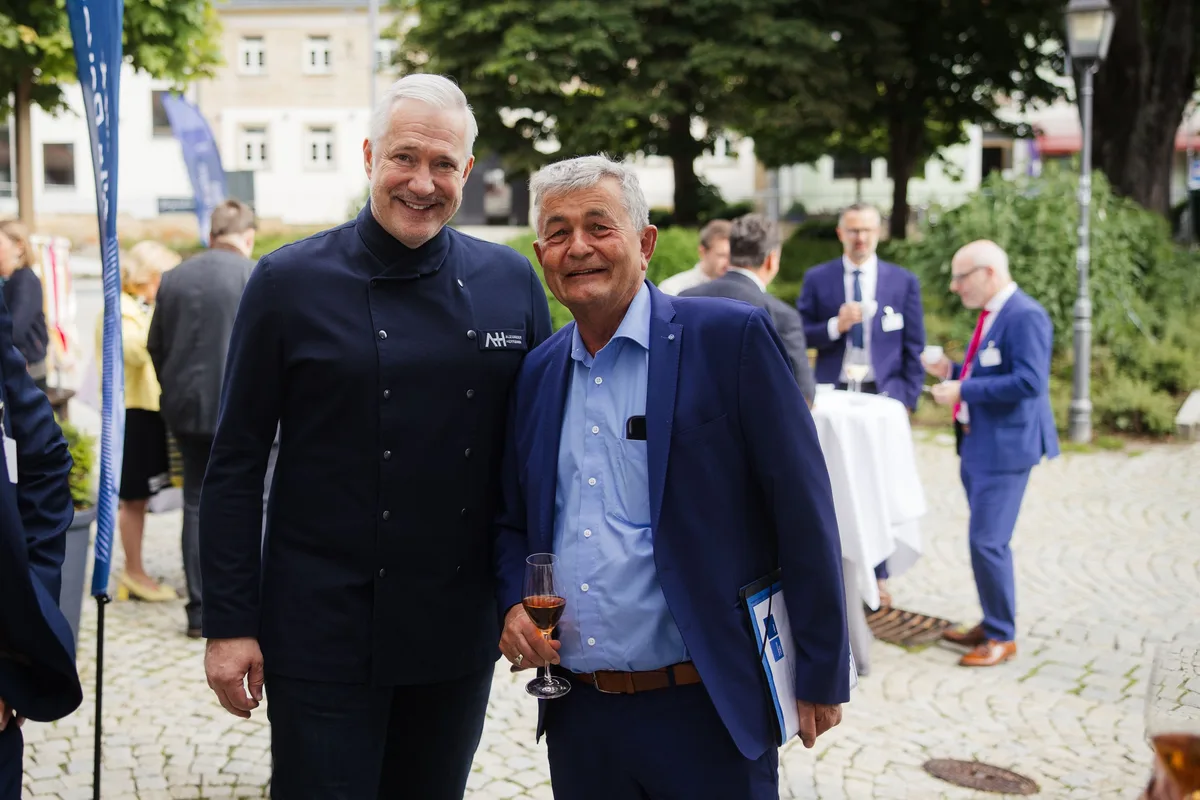 Koch in dunkler Kochjacke und älterer Mann im blauen Sakko mit Glas und Mappe lächeln nebeneinander in die Kamera