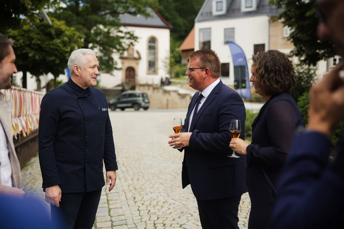 Koch im Gespräch mit einem Mann im dunklen Anzug und einer Frau, beide halten Gläser, dahinter Kopfsteinpflaster