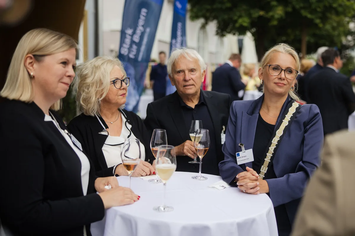 Vier Gäste mit Namensschildern und Gläsern am Stehtisch blicken zur Seite, hinter ihnen blaue Fahnen mit Löwen-Motiv