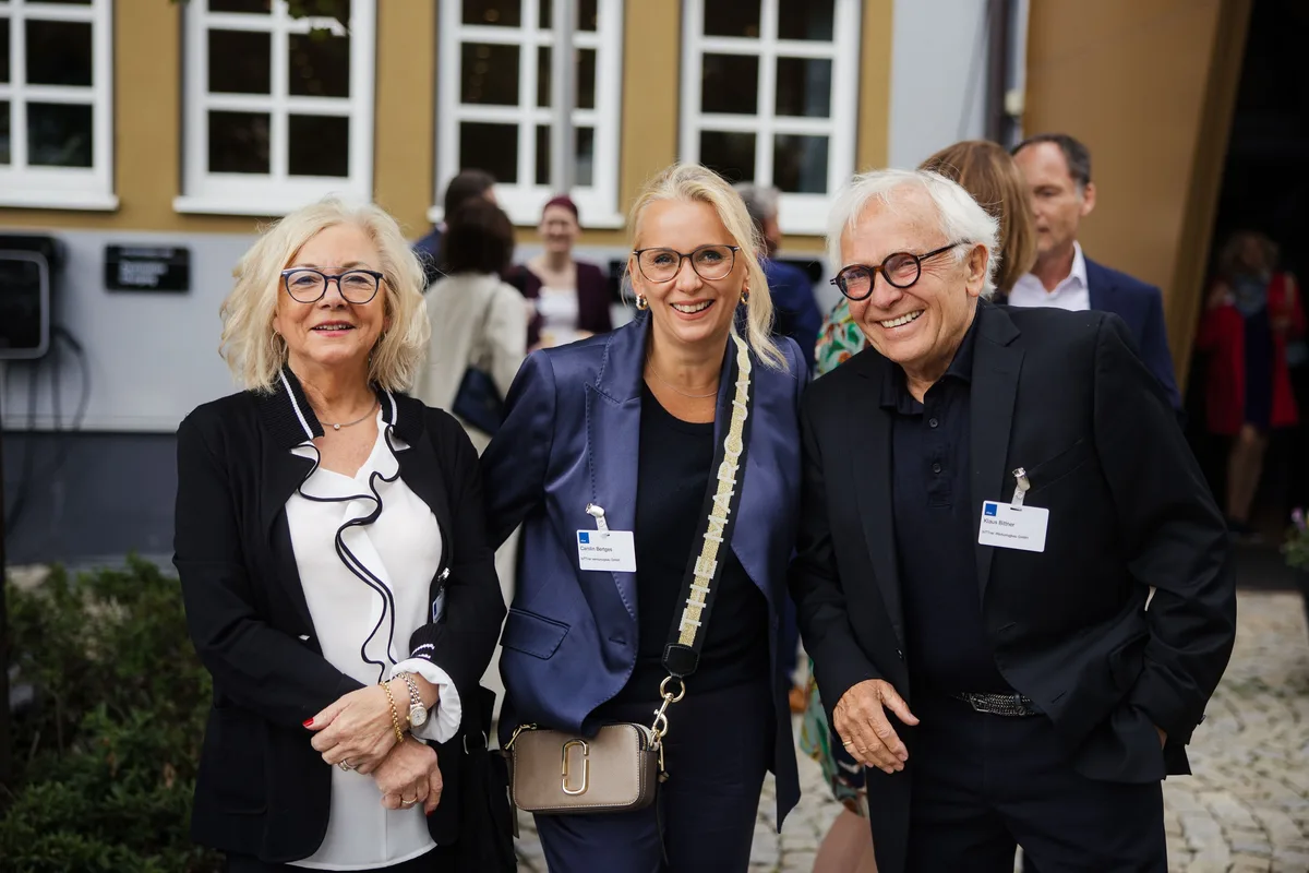 Drei Gäste lächeln eng zusammenstehend in die Kamera, zwei Frauen mit Brille und ein älterer Mann in Schwarz mit Namensschild