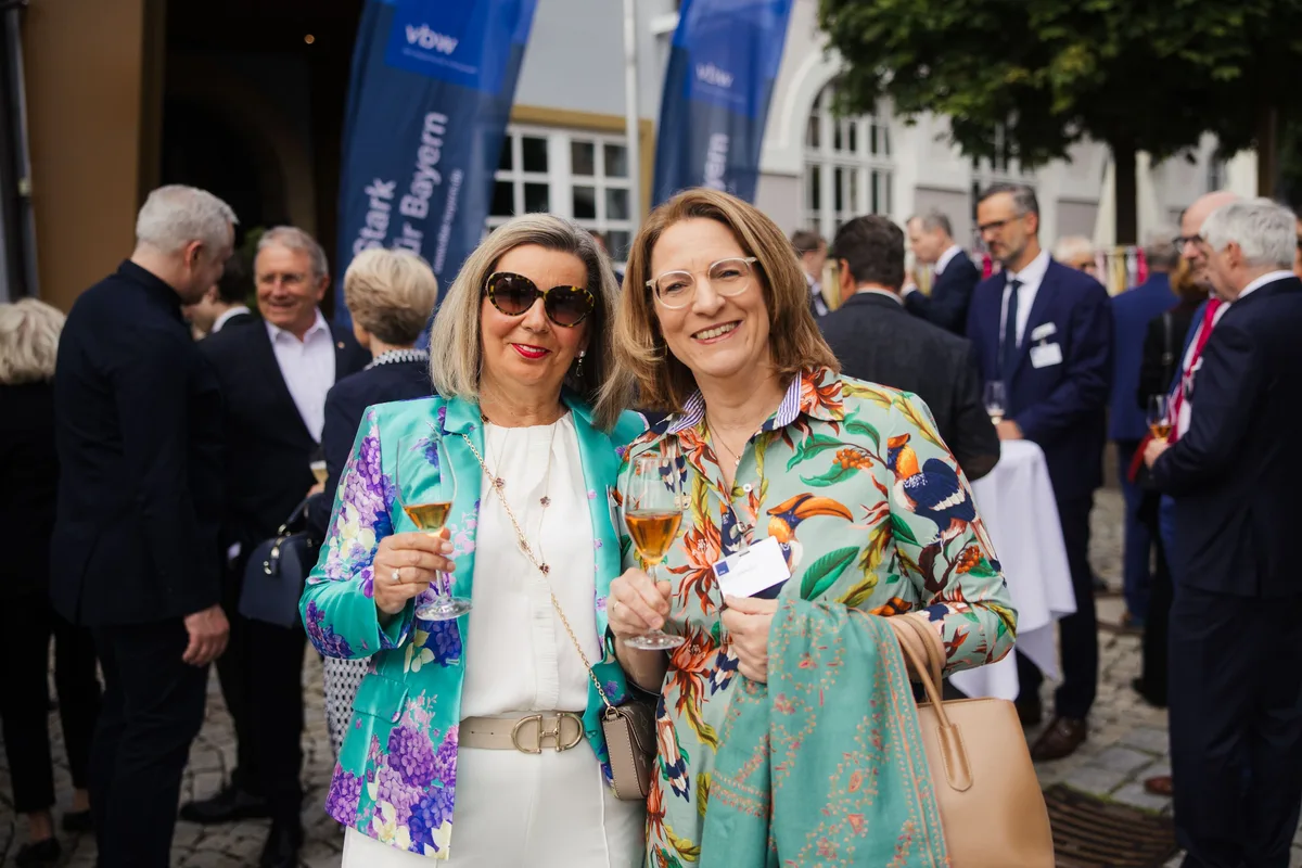 Zwei Frauen mit Gläsern lachen in die Kamera, eine in türkisfarbener Blumenjacke, die andere im Papageienkleid