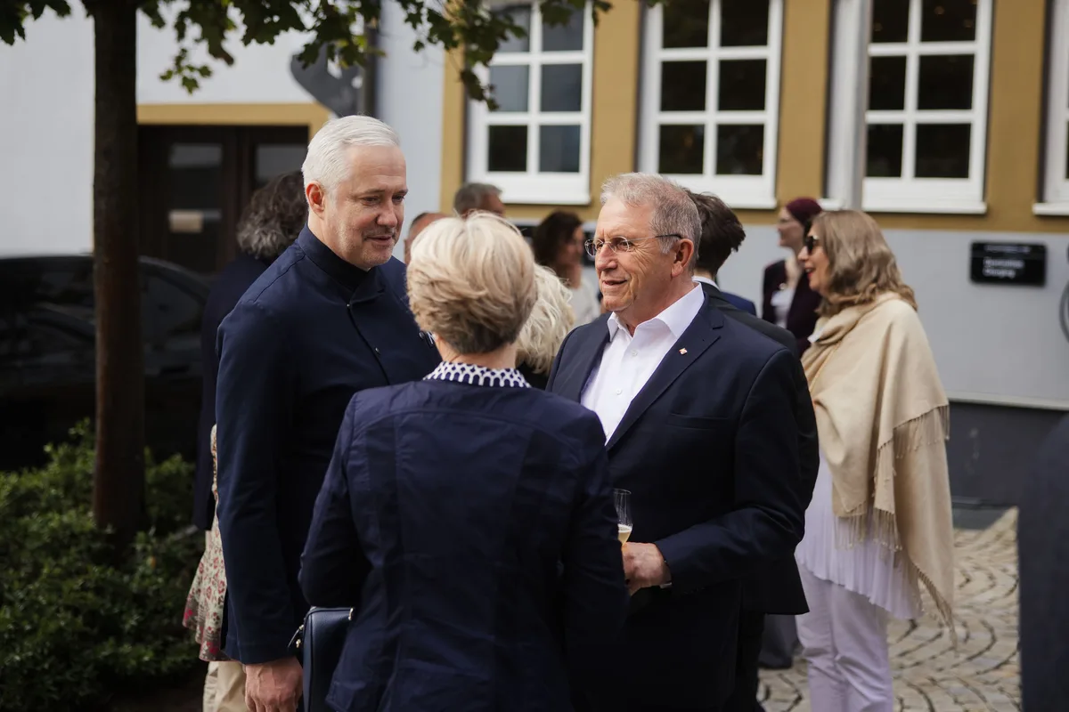 Koch im Gespräch mit einem älteren Mann mit Brille, davor eine Frau im dunklen Blazer von hinten, rechts eine Frau mit Schal