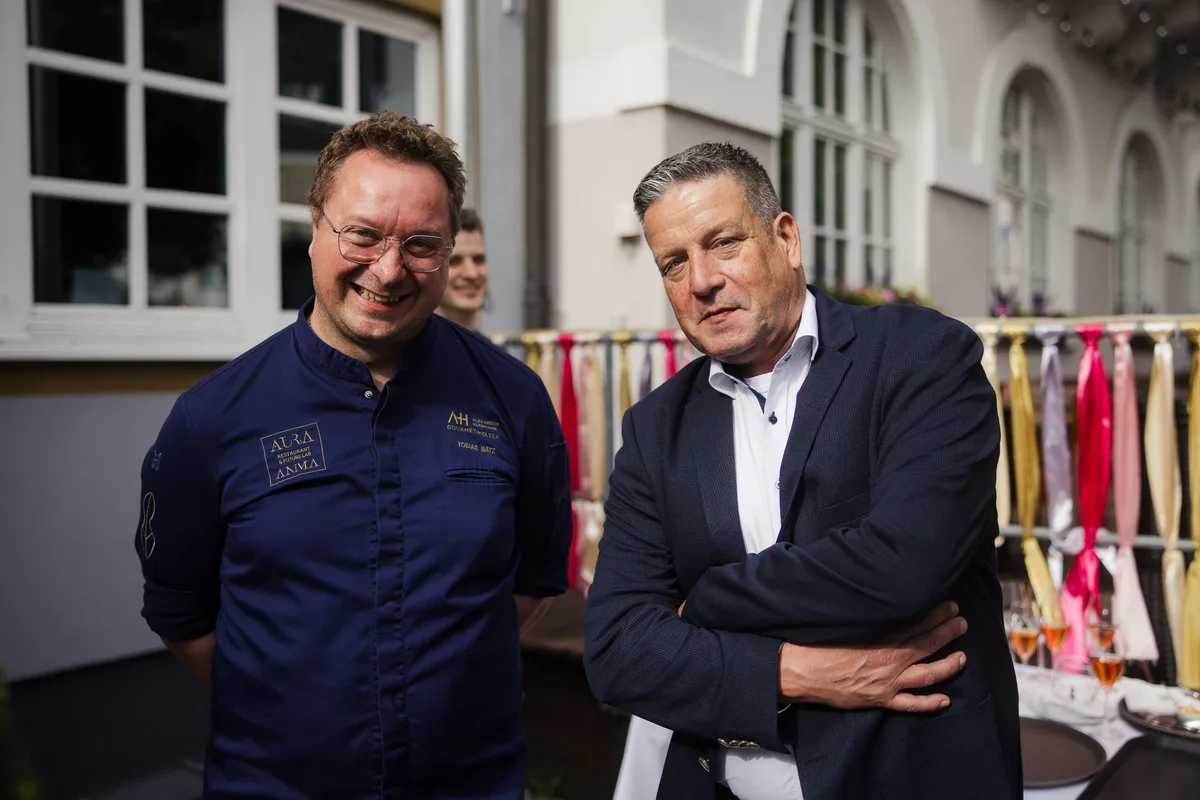 Lachender Koch mit Brille in blauer Jacke neben einem Mann im dunklen Sakko mit verschränkten Armen, dahinter Gläser