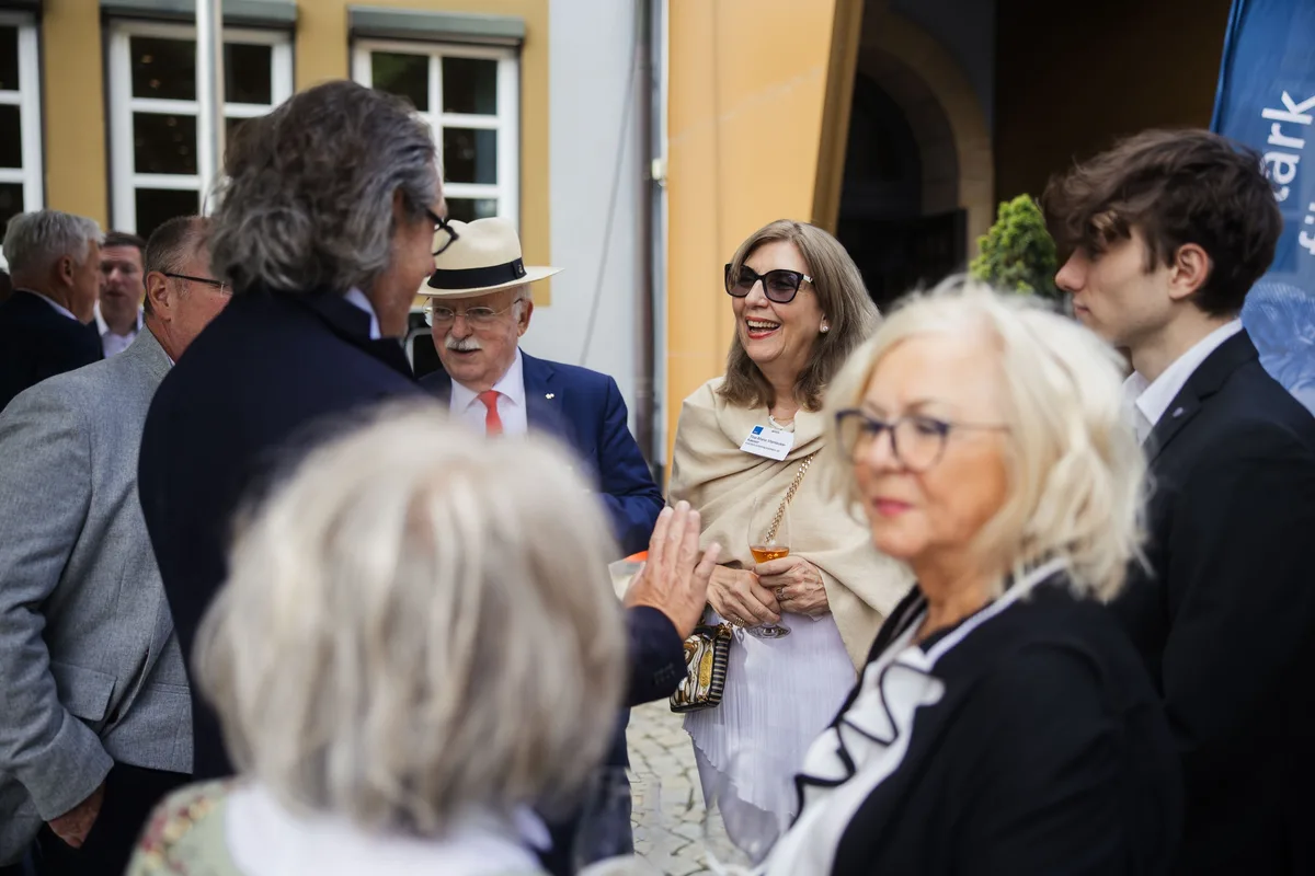 Frau mit Sonnenbrille und Schal lacht in einer dichten Gruppe, in der Mitte ein Mann mit Strohhut und Fliege