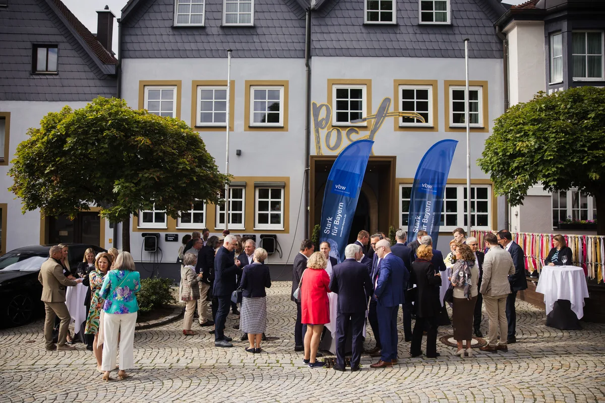 Weitwinkel auf den Vorplatz des Posthotels, Gäste in Gruppen an Stehtischen zwischen zwei Bäumen und blauen Fahnen