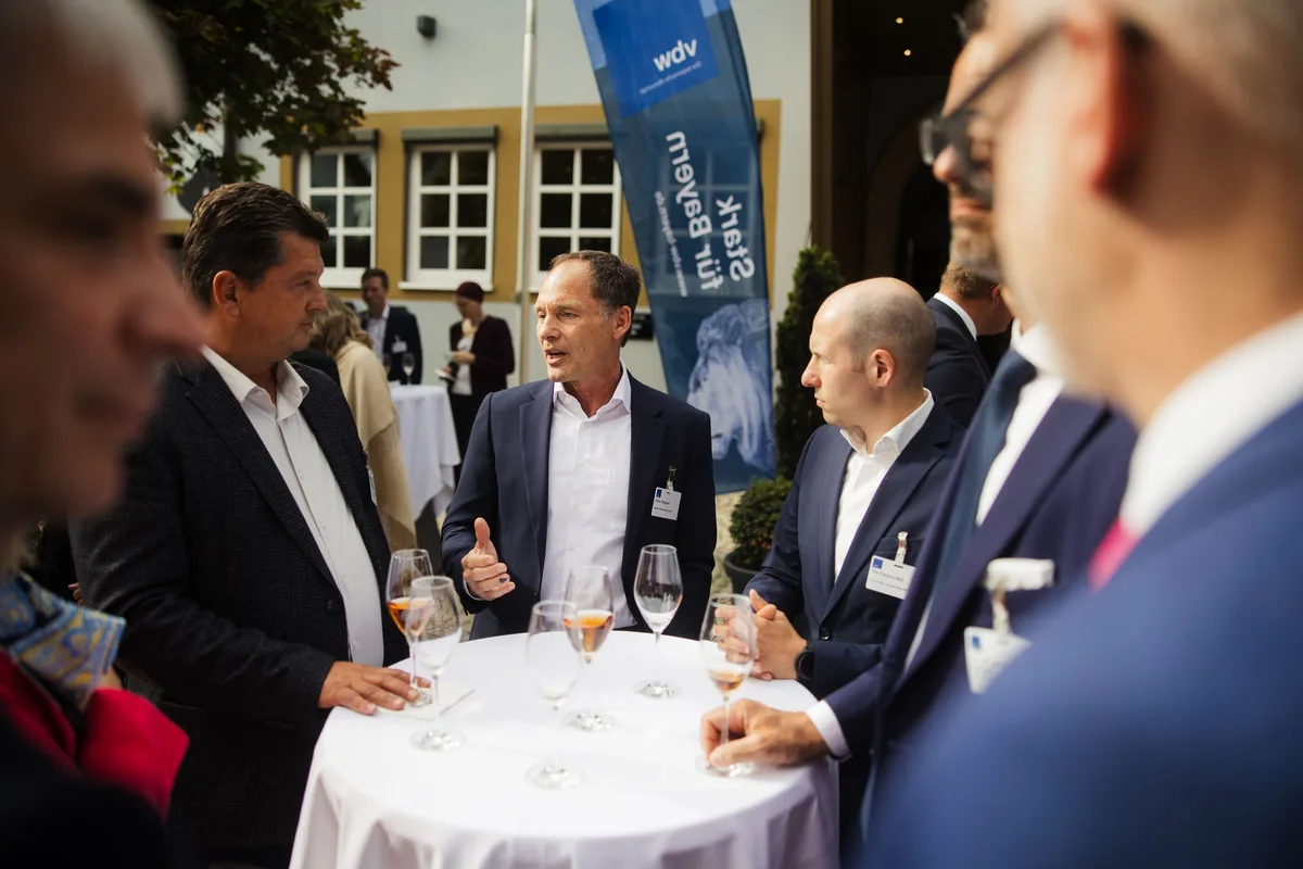 Vier Männer um einen Stehtisch mit Gläsern, einer erklärt mit erhobener Hand, links und rechts unscharfe Gesichter