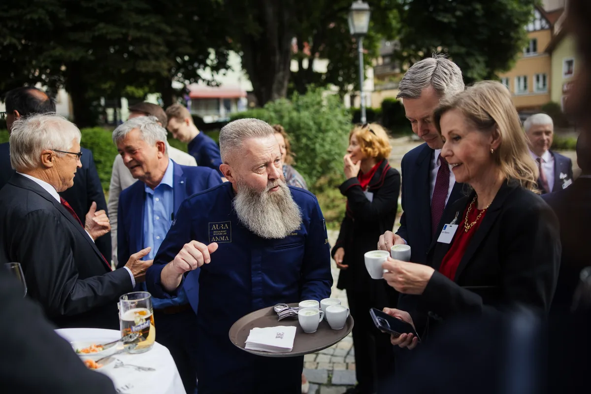 Bärtiger Koch hält ein Tablett mit Espressotassen und erklärt mit erhobener Hand, rechts nehmen Gäste Tassen