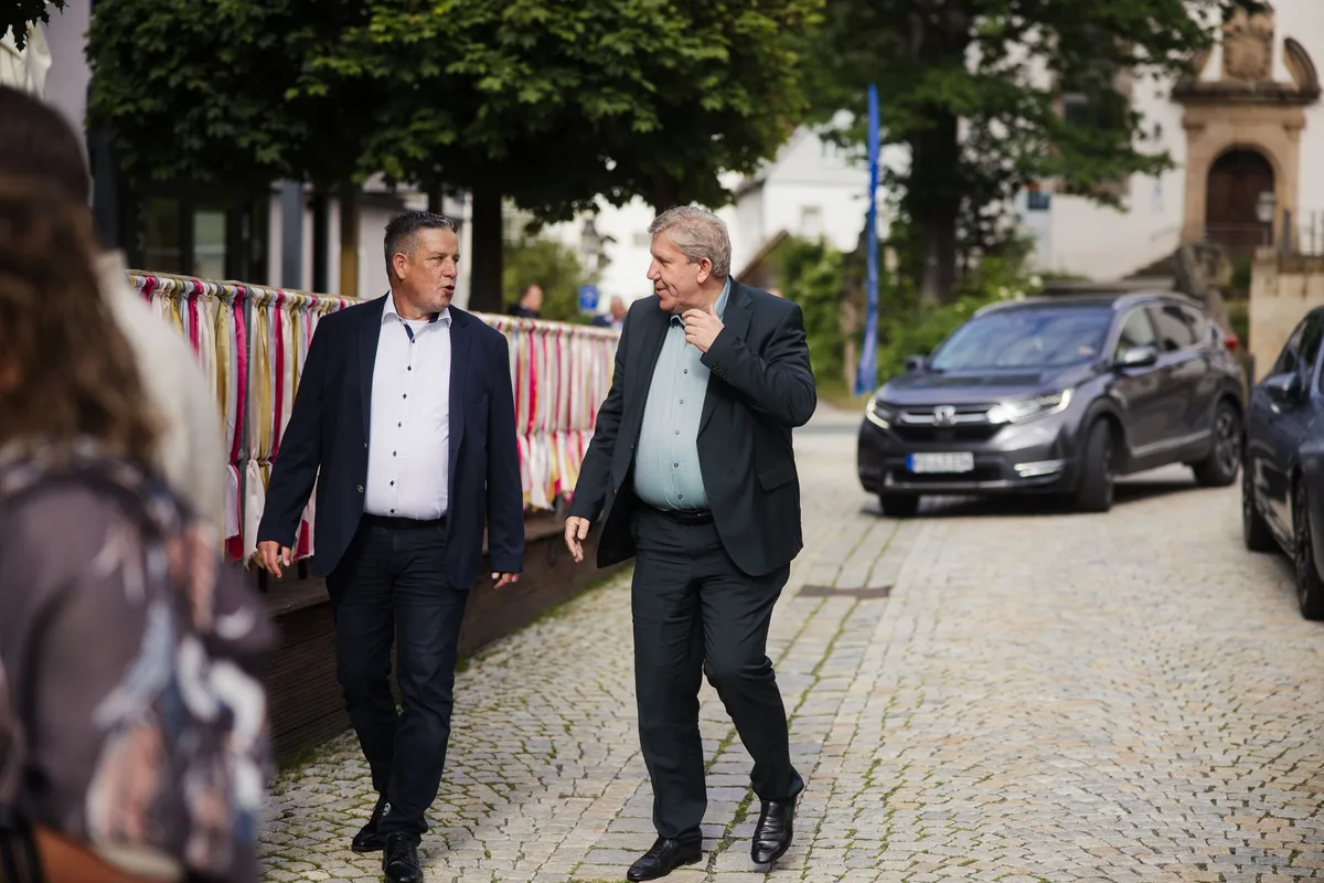 Zwei Männer in Anzug und Sakko gehen im Gespräch über das Kopfsteinpflaster, hinter ihnen parken Autos