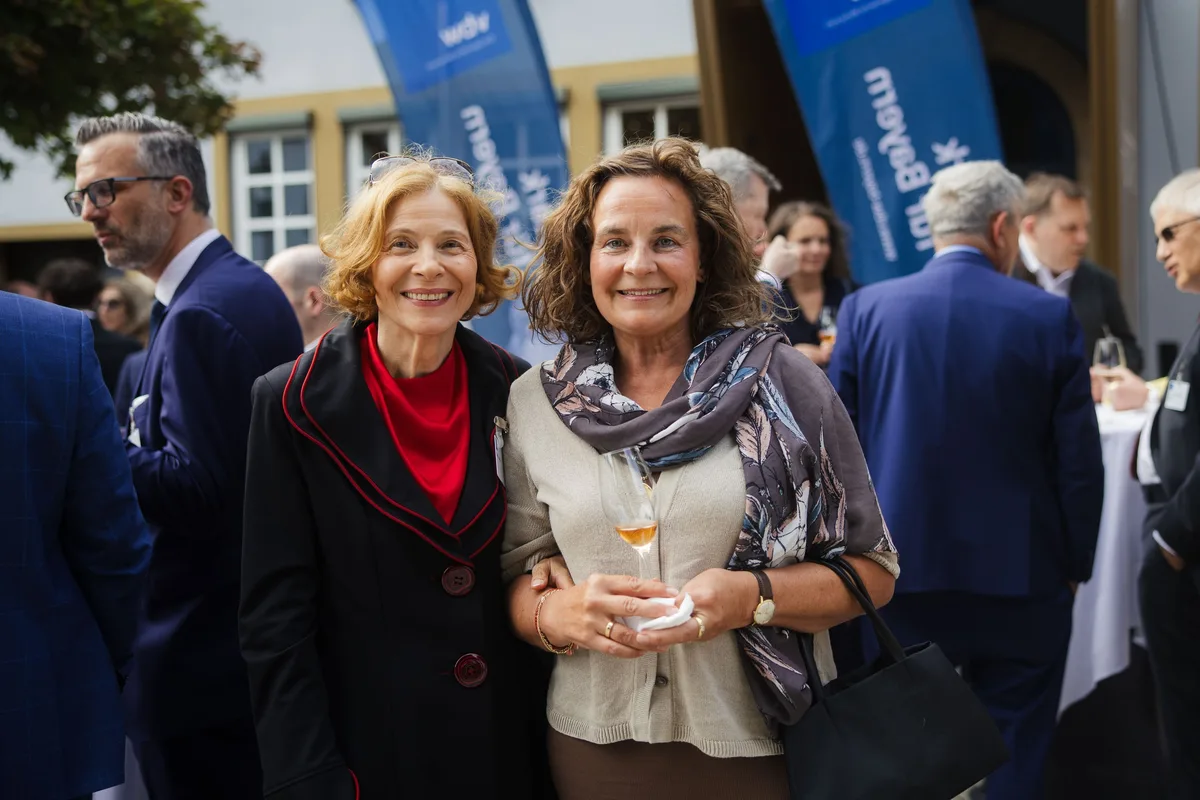 Zwei lächelnde Frauen stehen Arm in Arm, eine mit rotem Schal unter dem Jackett, die andere mit Mustertuch und Glas