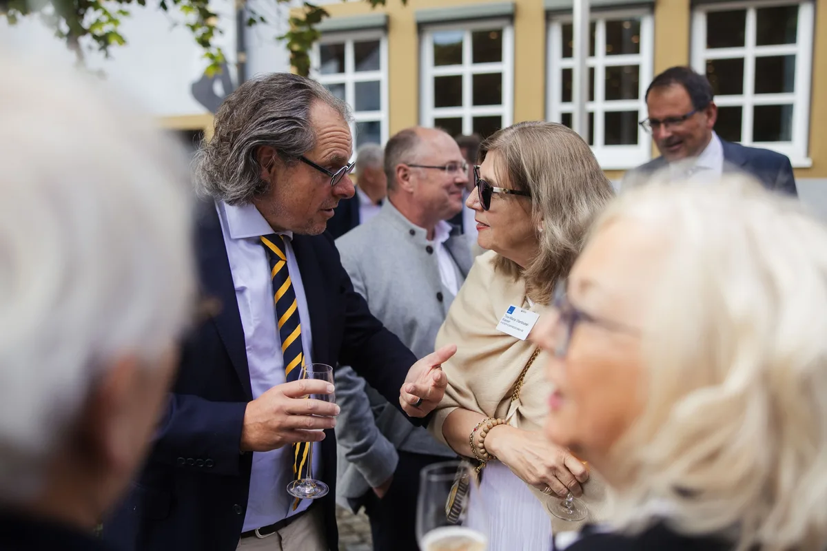 Mann mit langen grauen Haaren und gestreifter Krawatte gestikuliert im Gespräch mit einer Frau mit Sonnenbrille und Schal