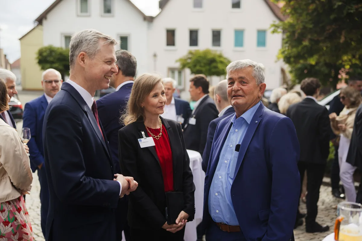 Mann im Anzug lacht im Gespräch mit einer Frau mit rotem Oberteil, rechts ein älterer Mann im blauen Sakko