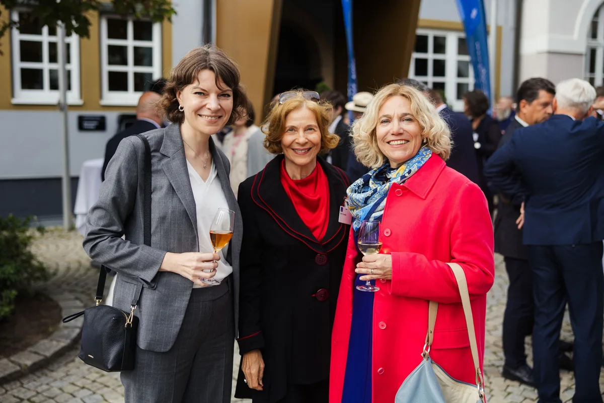 Drei Frauen mit Gläsern lächeln in die Kamera, die mittlere mit rotem Tuch, die rechte in roter Jacke, dahinter Gäste