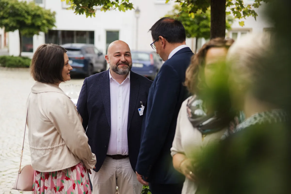 Lächelnder Mann mit Glatze und Bart im Gespräch mit einer Frau in heller Jacke, daneben ein Mann im Anzug von hinten