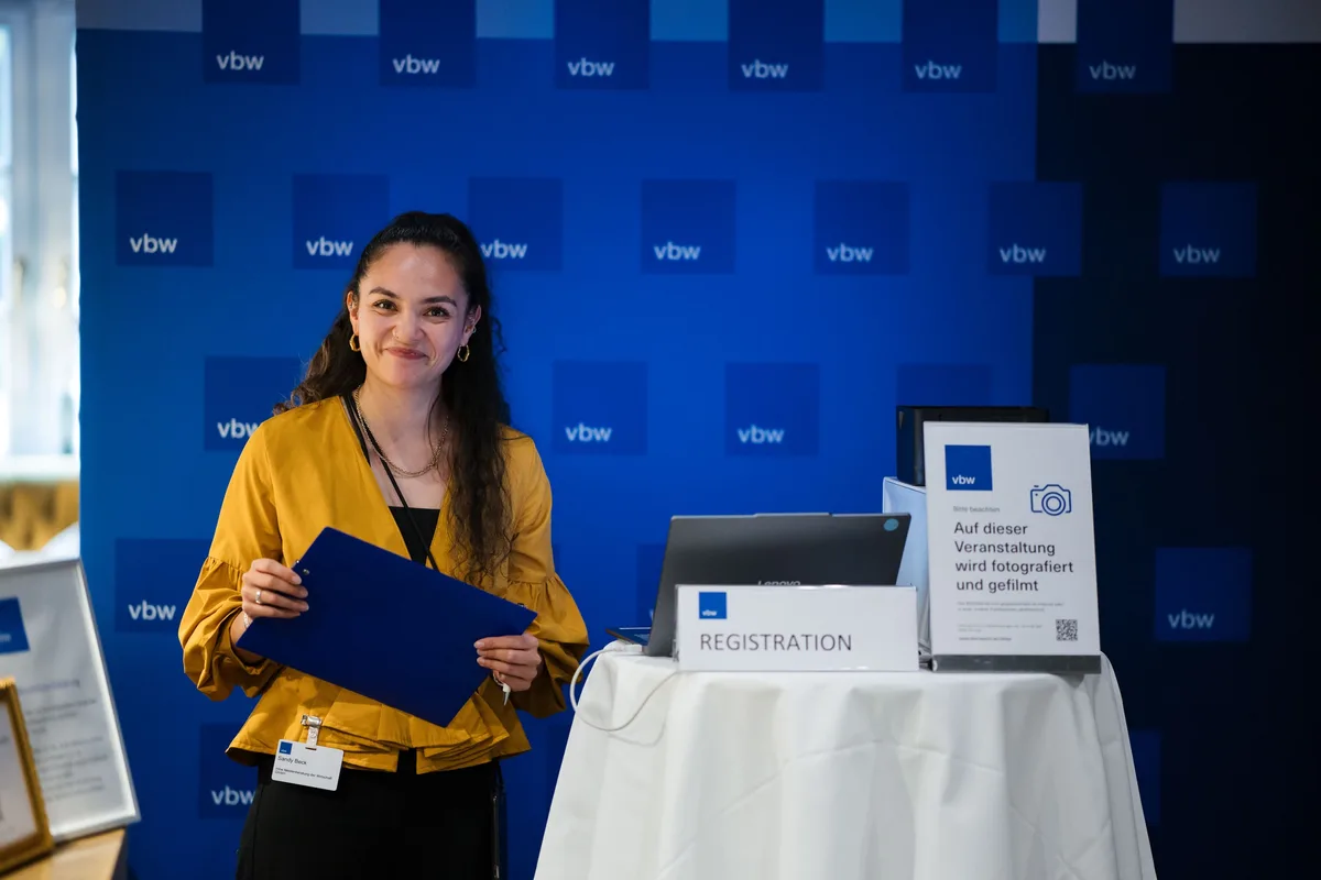 Junge Frau in gelber Bluse hält eine blaue Mappe am Empfangstisch vor der vbw-Wand, daneben ein Hinweisschild