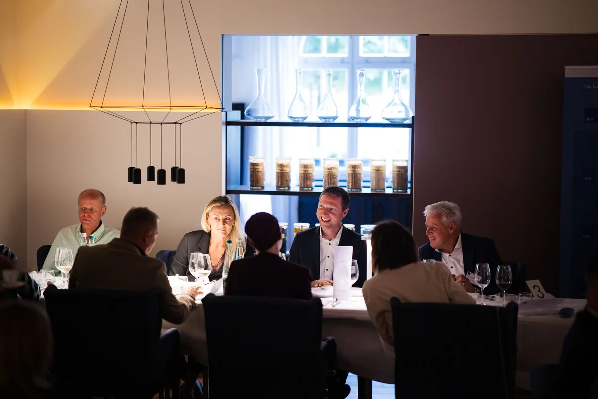 Gäste sitzen im abgedunkelten Saal an der Tafel, dahinter ein Regal mit Karaffen und Gläsern, darüber eine Pendelleuchte