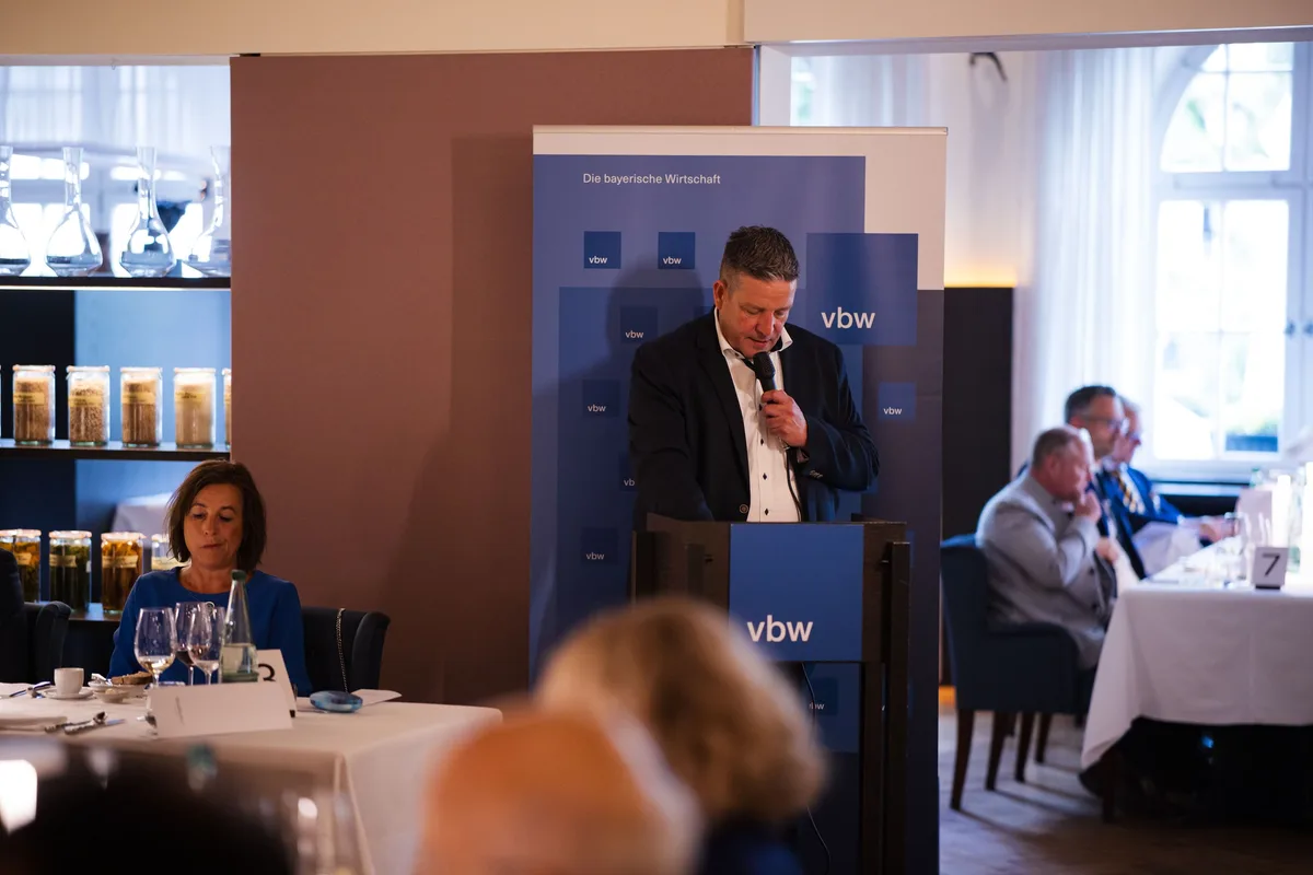 Redner am Pult mit Mikrofon vor einem vbw-Aufsteller, links im Vordergrund eine Frau in Blau am Tisch, rechts weitere Gäste