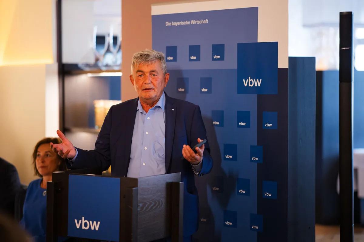 Älterer Redner im blauen Sakko hebt am vbw-Pult beide Hände, in einer Hand eine Fernbedienung, links im Anschnitt eine Frau