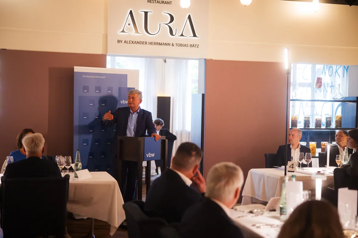 Redner am Pult unter dem AURA-Schriftzug, im Vordergrund Gäste von hinten, rechts Tische mit Gläsern und Tischnummer 4