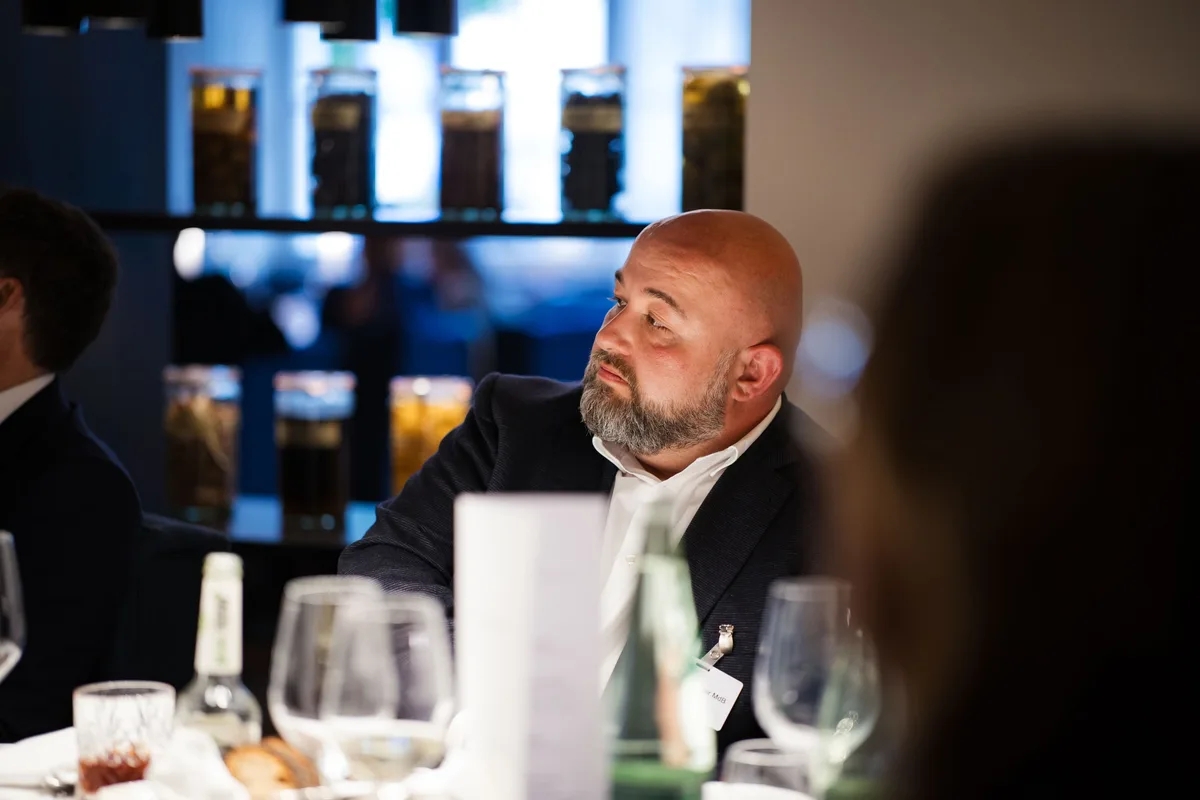 Mann mit Glatze und Bart blickt am Tisch seitlich aus dem Bild, hinter ihm ein Regal mit beleuchteten Gläsern