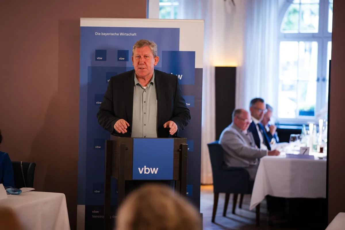 Redner im dunklen Sakko und grünem Hemd hinter dem vbw-Pult, rechts sitzen Gäste an einem Tisch vor dem Rundbogenfenster