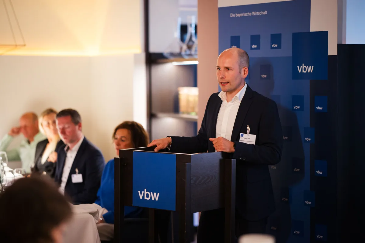 Glatzköpfiger Redner im Anzug stützt sich auf das vbw-Pult, links hören mehrere Gäste an der Tafel zu