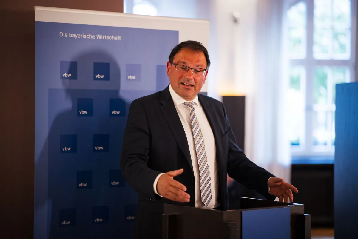 Redner mit Brille und gestreifter Krawatte öffnet am Pult beide Hände, hinter ihm die vbw-Wand und ein helles Fenster