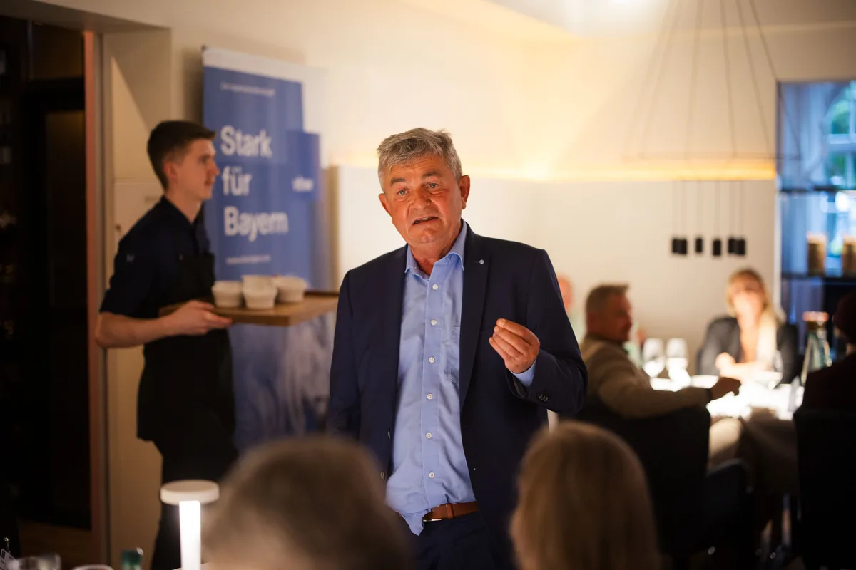 Redner im blauen Sakko steht im Saal, hinter ihm trägt ein Kellner ein Holzbrett mit Schälchen, rechts Gäste am Tisch