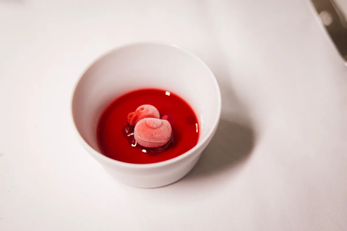 Kleine weiße Schale mit rotem Fruchtsud und zwei rosa Eiskugeln in Form von Schneckenhäuschen auf weißer Tischdecke