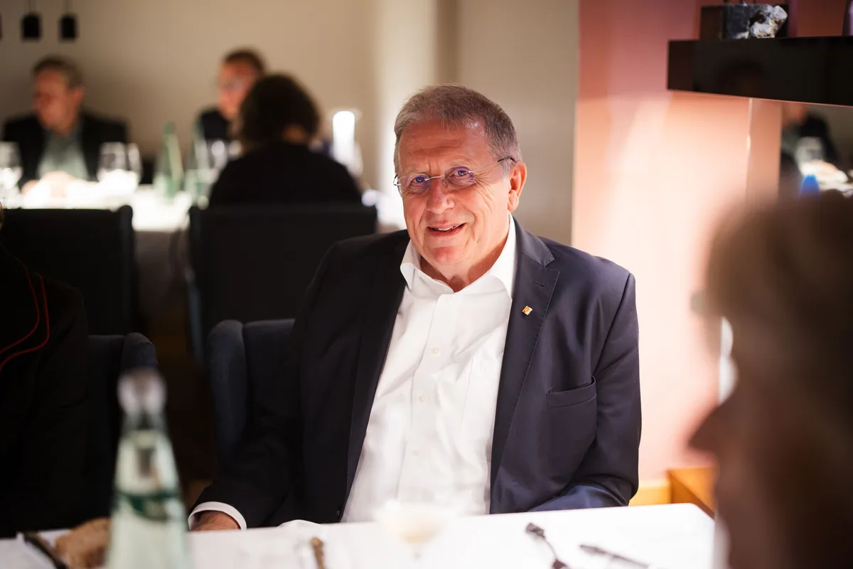 Älterer Mann mit Brille und weißem Hemd sitzt lächelnd am Tisch, im Hintergrund weitere Gäste im gedämpften Licht