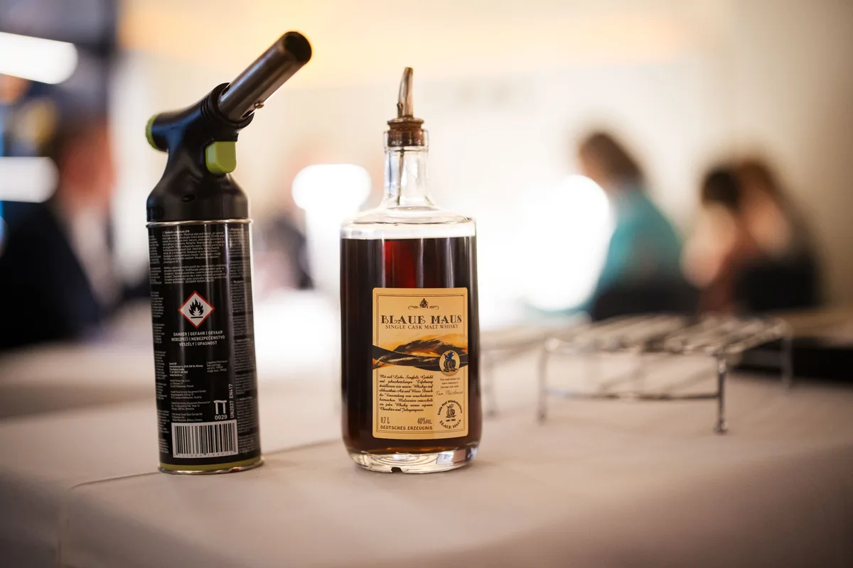 Brenner und Flasche Whisky auf dem Beistelltisch, dahinter verschwommen Gäste im hellen Saal