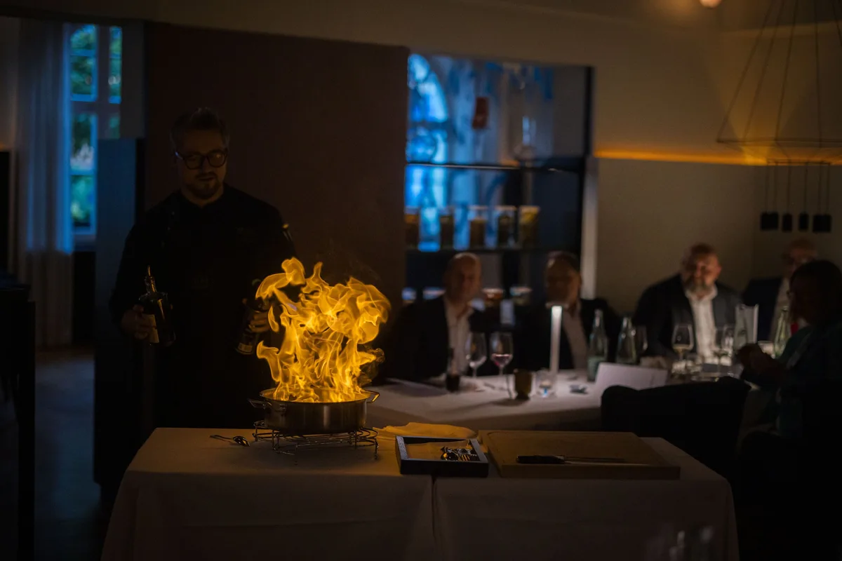 Koch flambiert in einem Topf mit hoher Flamme am Beistelltisch, im dunklen Saal sitzen Gäste im Hintergrund an der Tafel