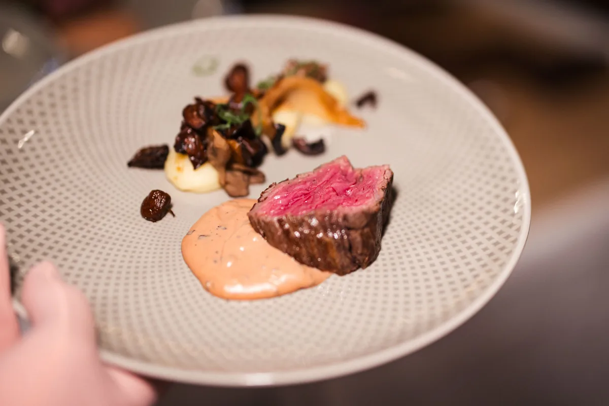 Hand hält einen gemusterten Teller mit rosa gebratenem Rindfleisch, Pilzen, Püree und heller Sauce