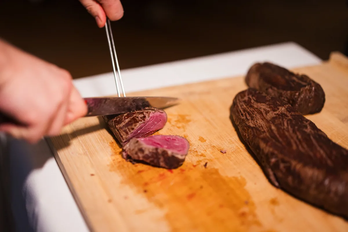 Messer und Gabel schneiden rosa gebratenes Fleisch auf einem Holzbrett, rechts liegt der restliche Braten