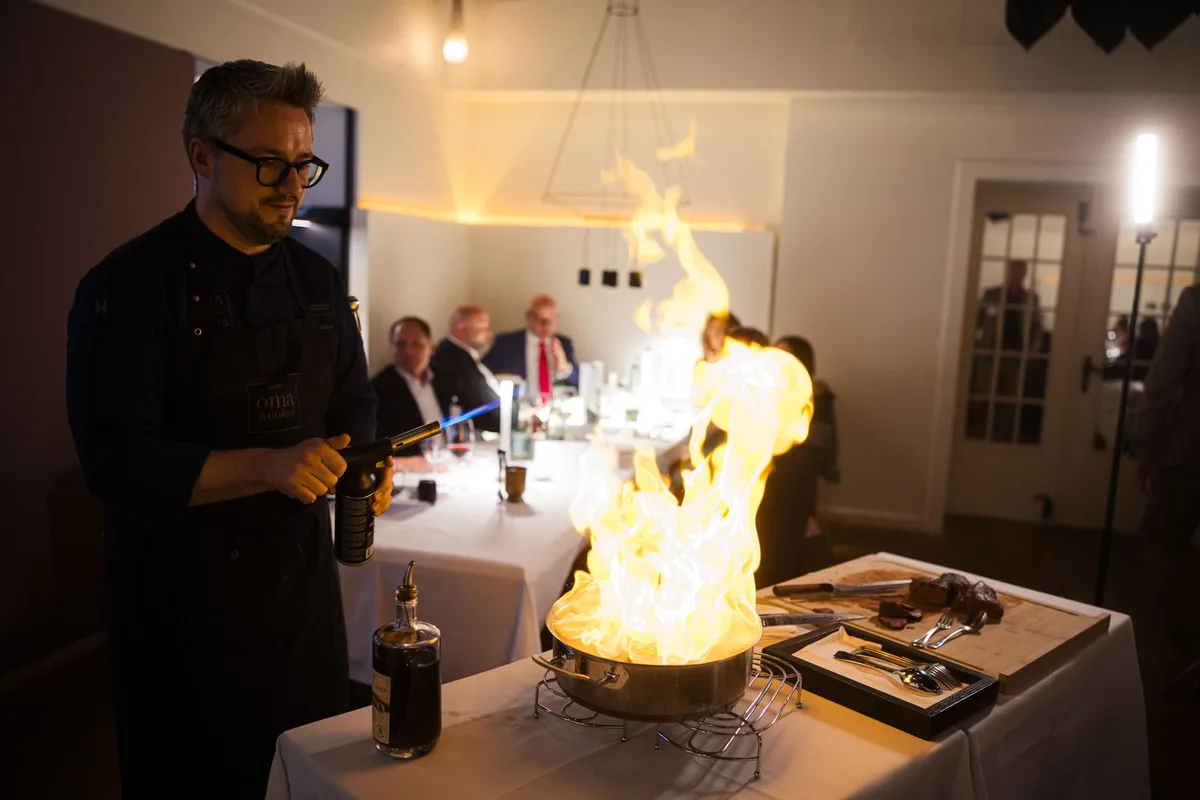 Koch mit Brille und Bart flambiert mit dem Brenner in einer Pfanne, im Hintergrund sitzen Gäste an der Tafel