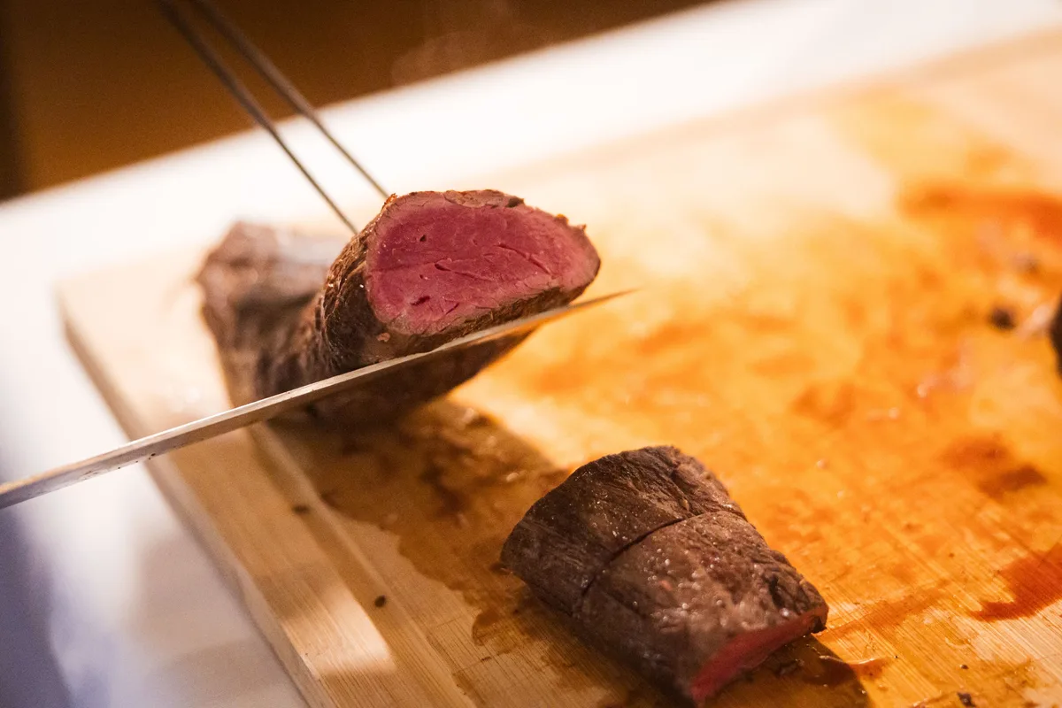 Aufgeschnittener Braten auf dem Holzbrett, eine lange Klinge hebt ein Stück an, das Fleisch ist innen rosa