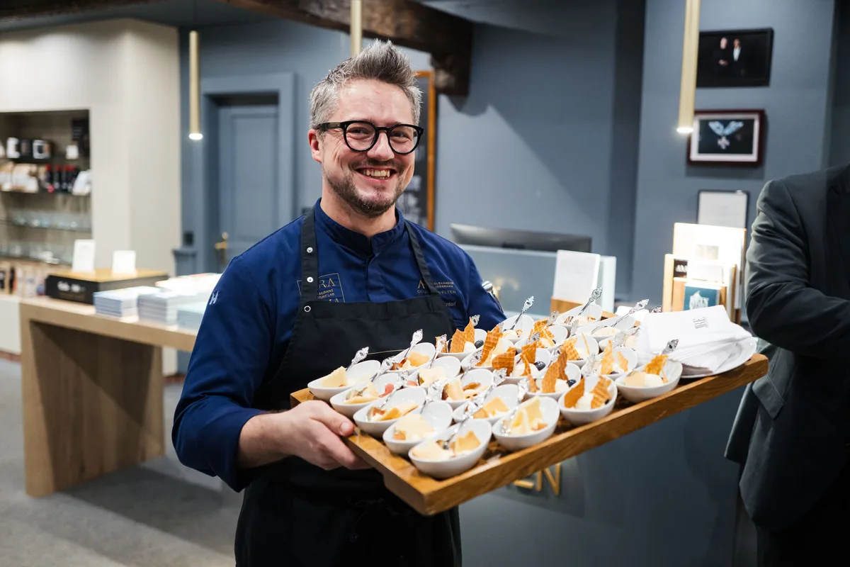Lächelnder Koch mit Brille und Schürze hält ein Holztablett mit Dessertschälchen in der Hotelhalle