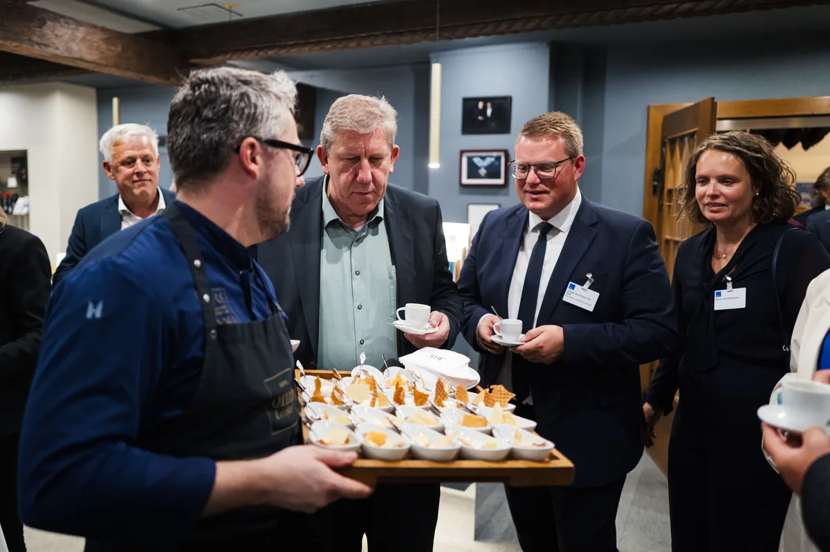 Koch mit Dessert-Tablett im Gespräch mit mehreren Gästen, die Kaffeetassen halten, rechts eine Frau mit Namensschild