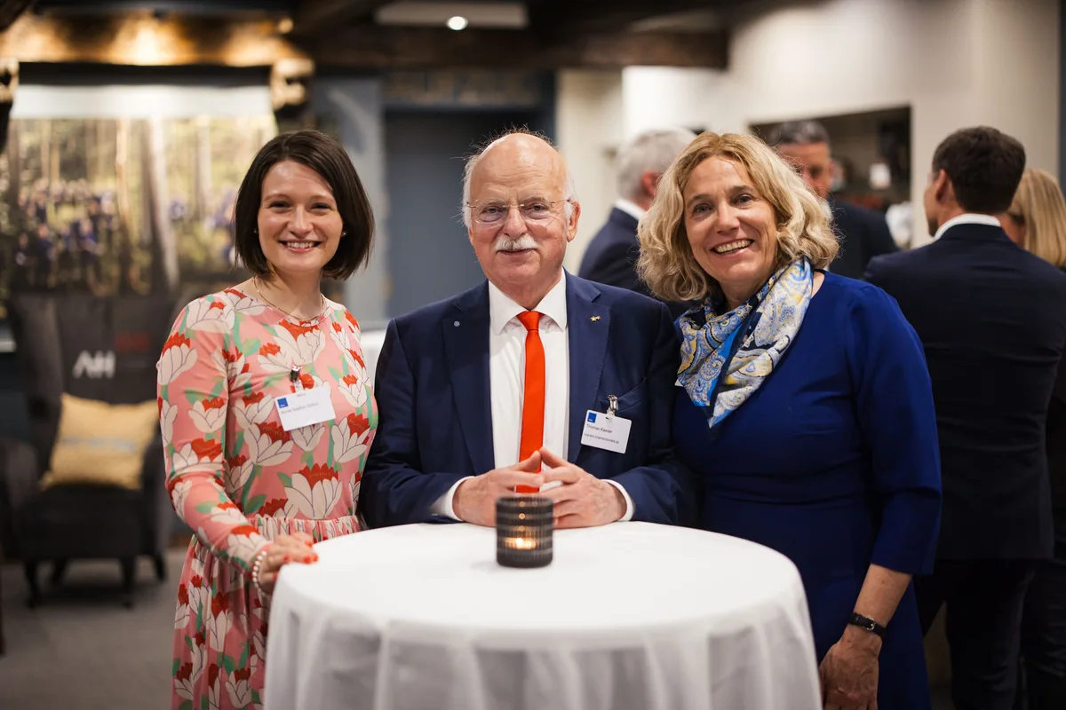 Zwei Frauen und ein älterer Mann mit orangefarbener Krawatte stehen lächelnd an einem Stehtisch mit Kerze