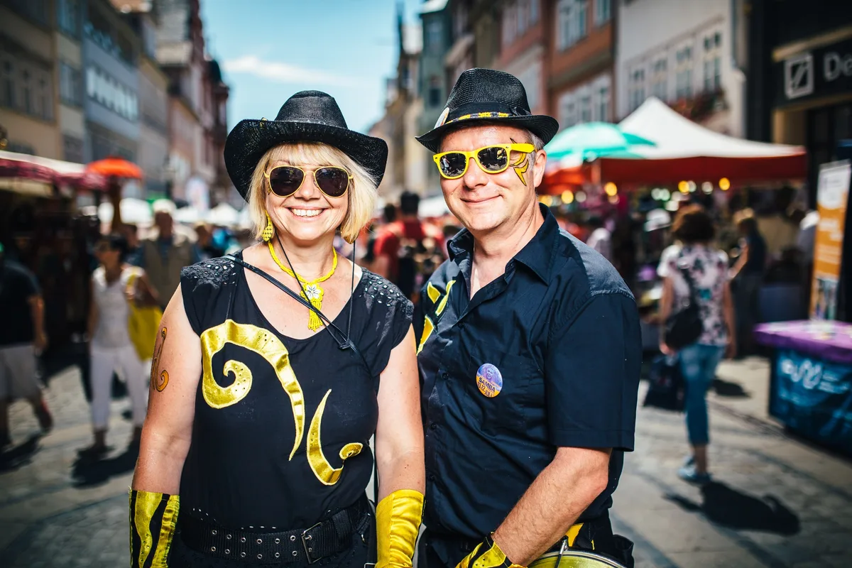 Lächelndes Paar in Schwarz mit goldenen Mustern, schwarzen Hüten und Sonnenbrillen auf einer Altstadtgasse